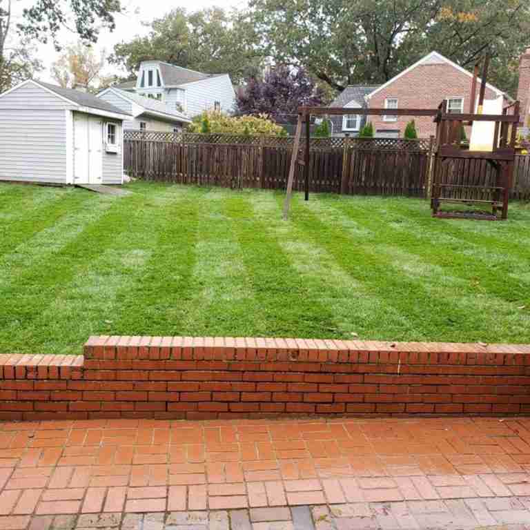 A backyard with a neatly mowed lawn featuring striped patterns, a brick patio and low wall, a small shed, and a wooden play structure.