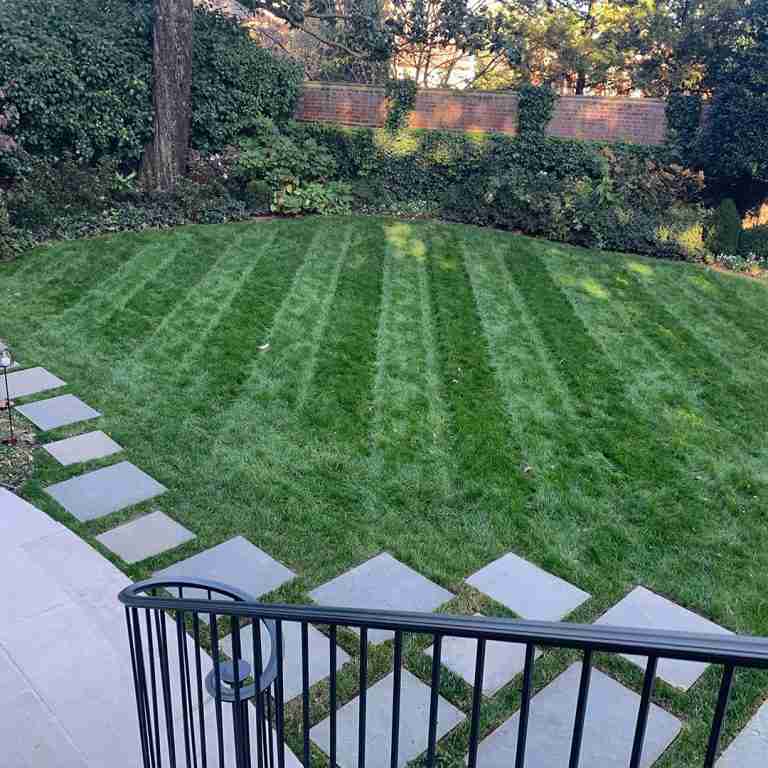 A neatly mowed lawn with distinct stripes, bordered by a brick wall and foliage, with a stone pathway leading from the foreground.