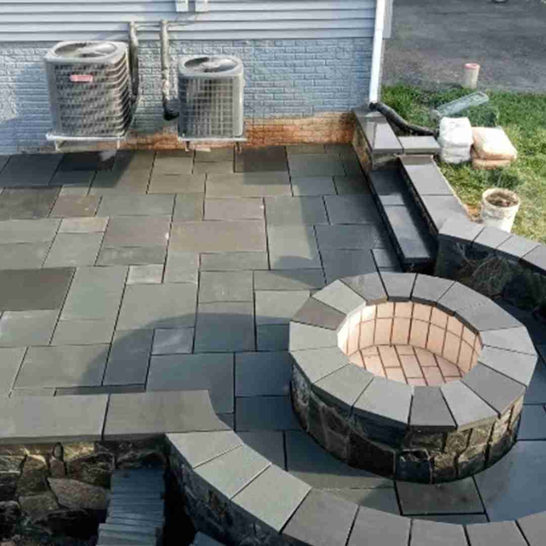 Outdoor patio with neatly arranged grey stone pavers and a circular stone fire pit. Two air conditioning units are mounted on the house wall nearby.
