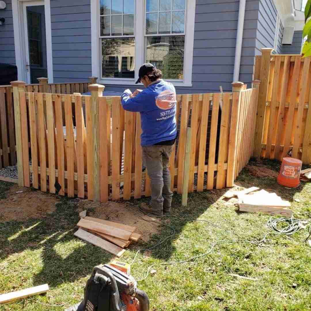 A person constructing a wooden fence in a backyard, wearing a blue shirt and cap, with tools and materials scattered on the grass.