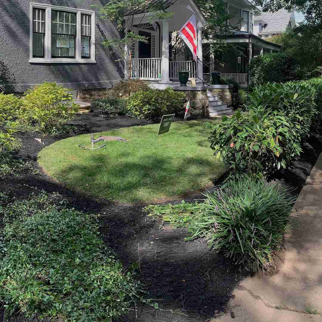 A front yard with a neatly maintained lawn, bordered by various shrubs and plants. A small sprinkler is watering the grass, and a pathway leads to a house with a porch. An American flag is displayed near the entrance.