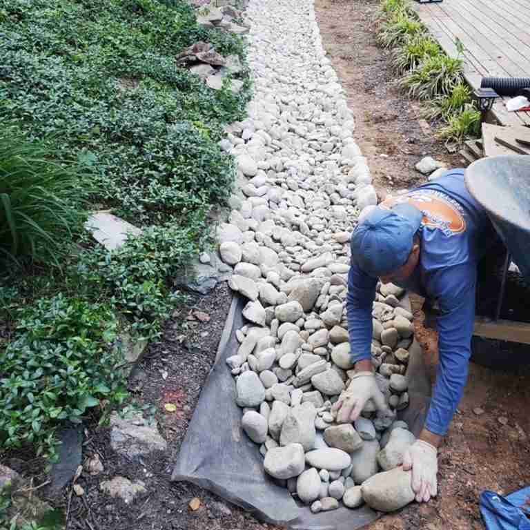 A person wearing gloves arranges stones on a fabric strip, creating a pathway, with greenery on one side and soil on the other.