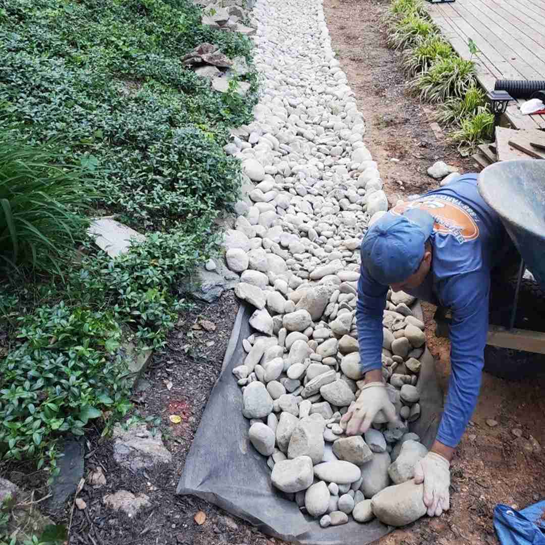A person wearing gloves arranges stones on a fabric strip, creating a pathway, with greenery on one side and soil on the other.