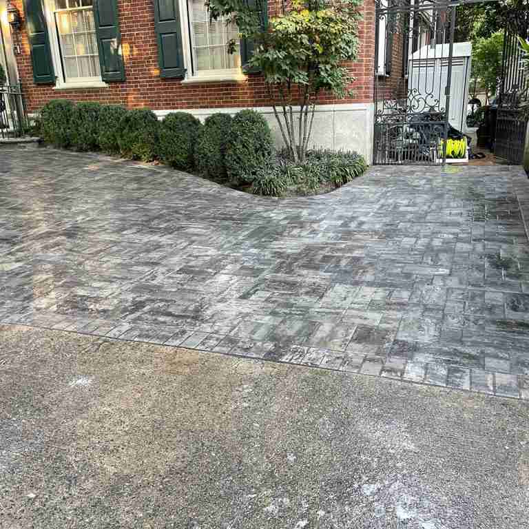 A paved driveway with a decorative pattern leads to a gated entrance next to a red brick building, lined with shrubs and a small tree.