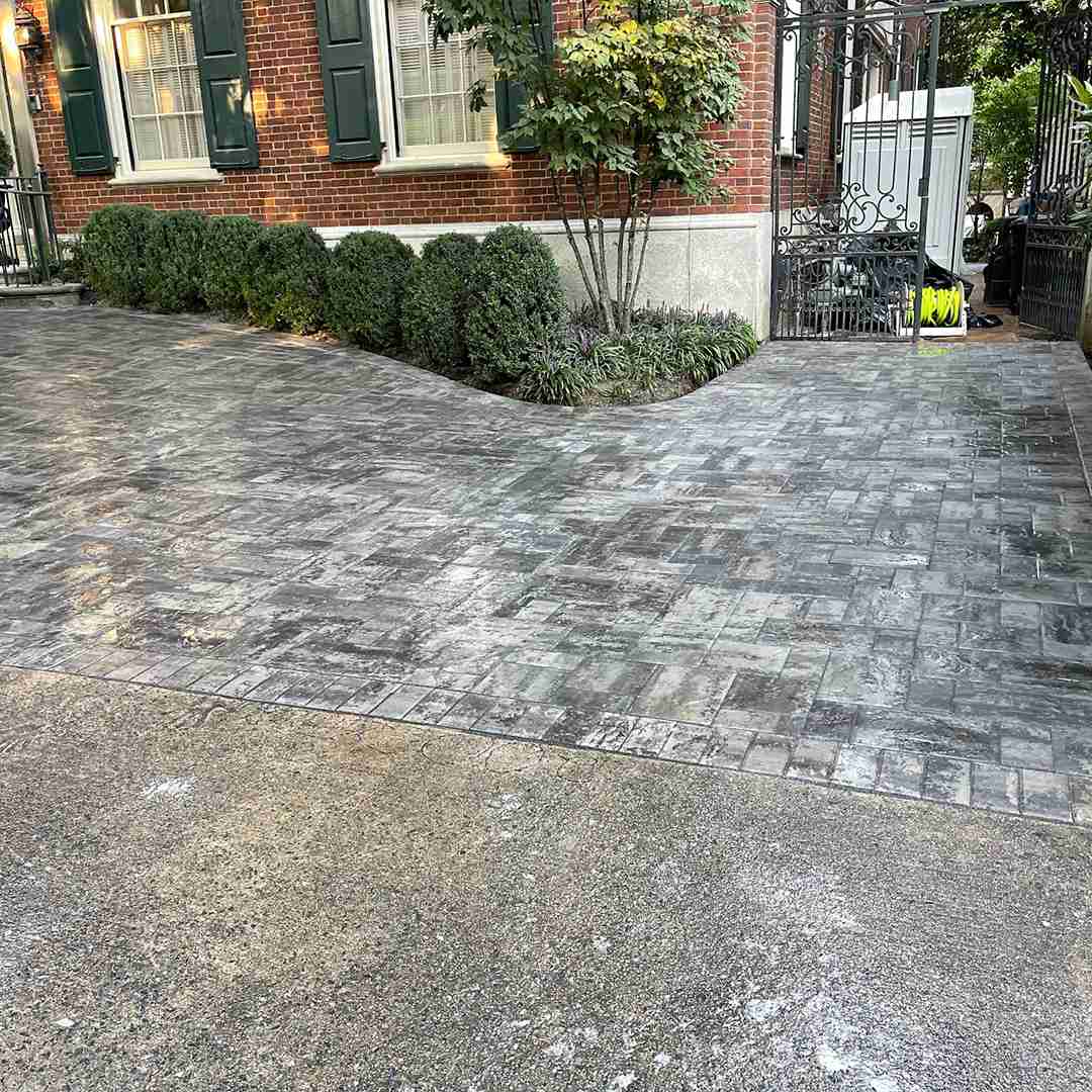 A paved driveway with a decorative pattern leads to a gated entrance next to a red brick building, lined with shrubs and a small tree.