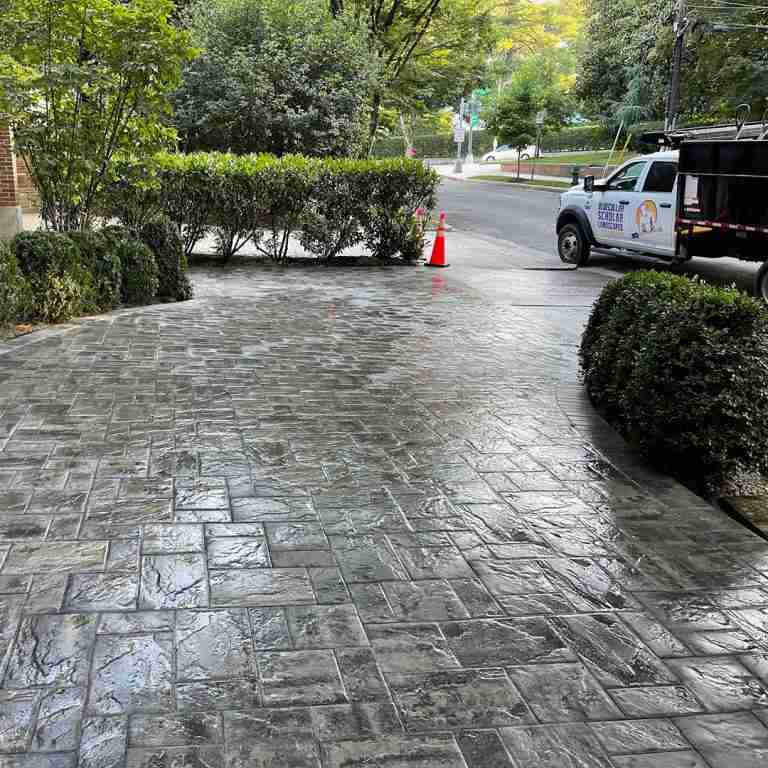 Freshly sealed driveway with decorative stone pattern, bordered by hedges, with a work truck and traffic cones in the background.