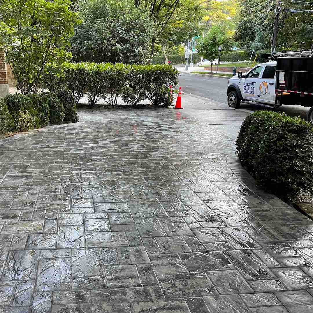 Freshly sealed driveway with decorative stone pattern, bordered by hedges, with a work truck and traffic cones in the background.