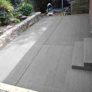 A smooth, freshly poured concrete driveway with grooves, surrounded by a stone retaining wall. A person is working with a power tool in the background.
