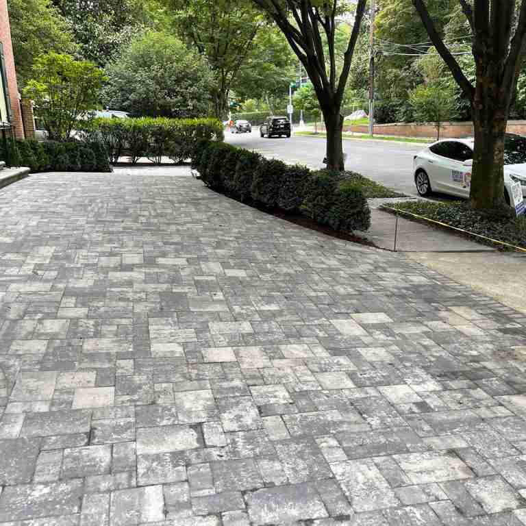A paved driveway with interlocking stones, bordered by neatly trimmed hedges and trees, leads to a street with parked and passing cars.