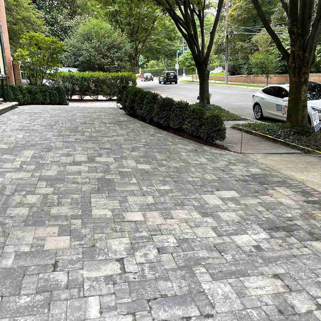 A paved driveway with interlocking stones, bordered by neatly trimmed hedges and trees, leads to a street with parked and passing cars.