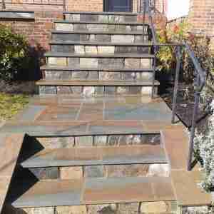 Stone steps with a metal handrail leading to an entrance, surrounded by brick walls and greenery.