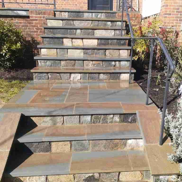Stone steps with a metal handrail leading to an entrance, surrounded by brick walls and greenery.