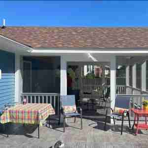 Patio with a table covered in a colorful checkered cloth, two chairs with floral cushions, and a small red table with a potted plant, all under a covered porch. A cat is lounging on the wooden deck.