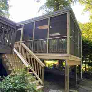 Screened-in porch with wooden railings and stairs, elevated on wooden posts, surrounded by trees.