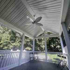 Covered porch with a ceiling fan, white railing, and folding chairs, overlooking a wooded area.