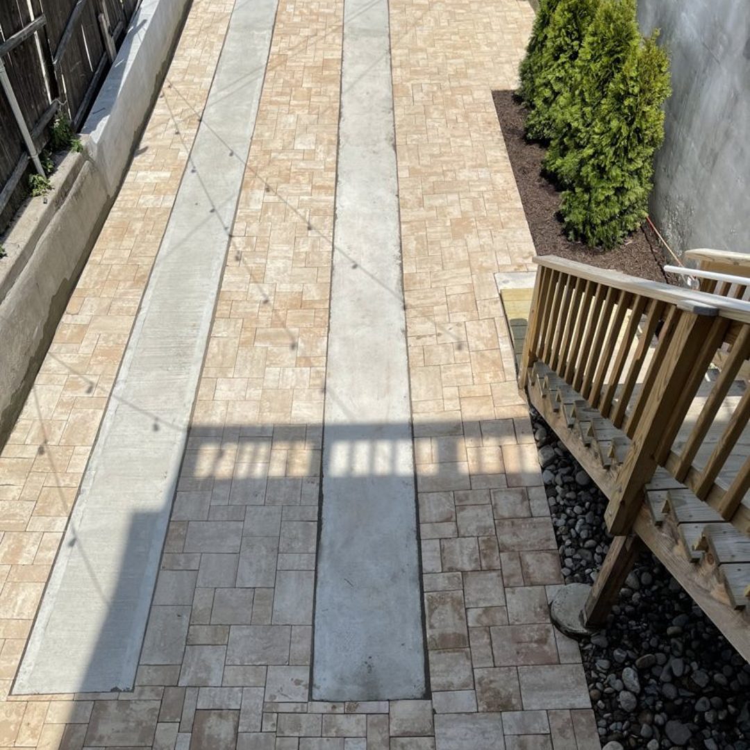 Paved backyard with two narrow concrete strips for vehicle tires, surrounded by brick pavers, wooden stairs on the right, and small shrubs planted beside a wall.