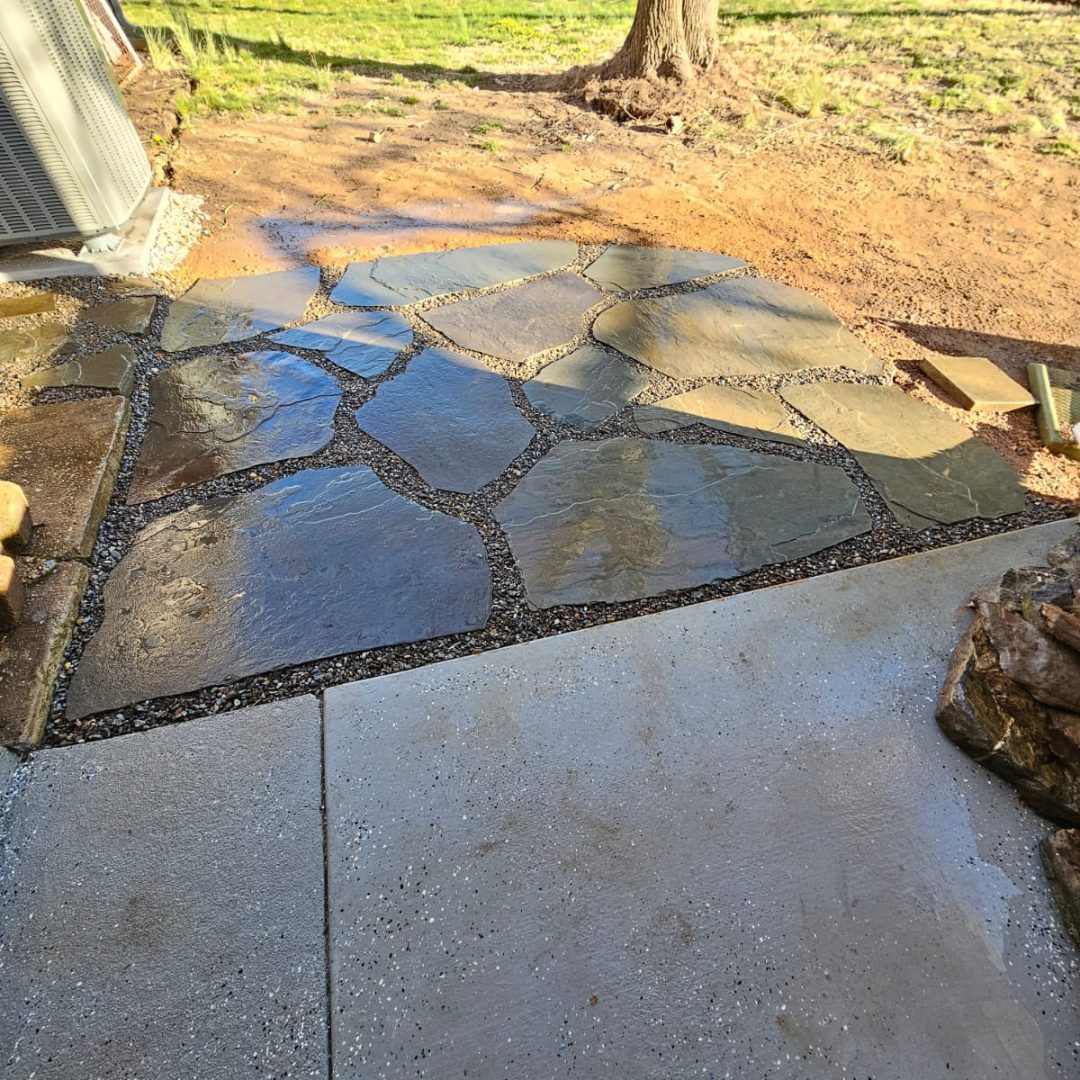 Wet irregular stone patio next to concrete slab, with grass and a tree in the background.