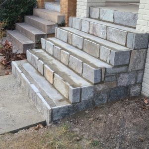 Stone staircase with four steps leading up to a brick building entrance. The steps are made of rectangular stone blocks and are adjacent to a concrete path.