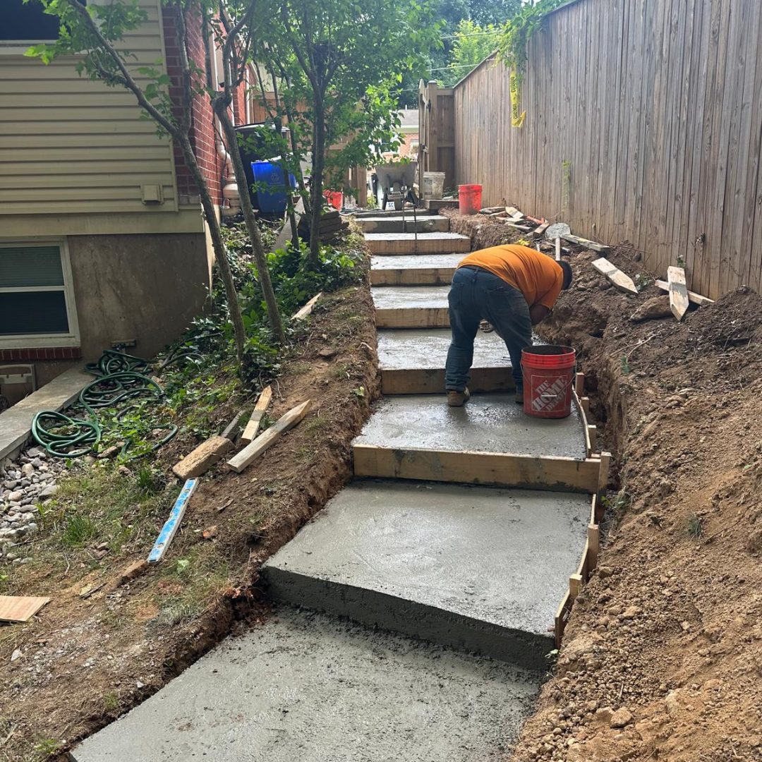 Worker constructing cement steps along a hillside next to a house and wooden fence.