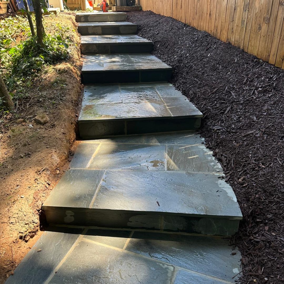 Stone steps leading up a slope, bordered by a wooden fence on one side and soil on the other.