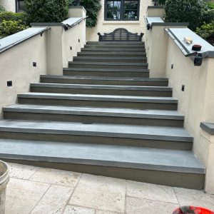 Wide stone steps leading up to a building entrance, flanked by beige walls and greenery.