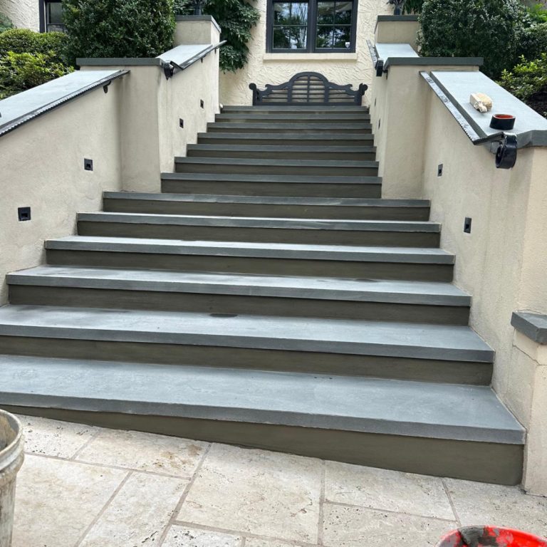 Wide stone steps leading up to a building entrance, flanked by beige walls and greenery.