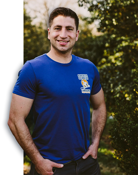 A man in a blue t-shirt with "Blue Collar Scholar Landscapes" logo stands smiling with hands in pockets, outdoors with greenery in the background.