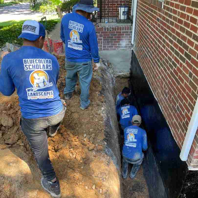 Workers from "Blue Collar Scholars Landscapes" wearing blue shirts are excavating and working on a foundation next to a brick building.