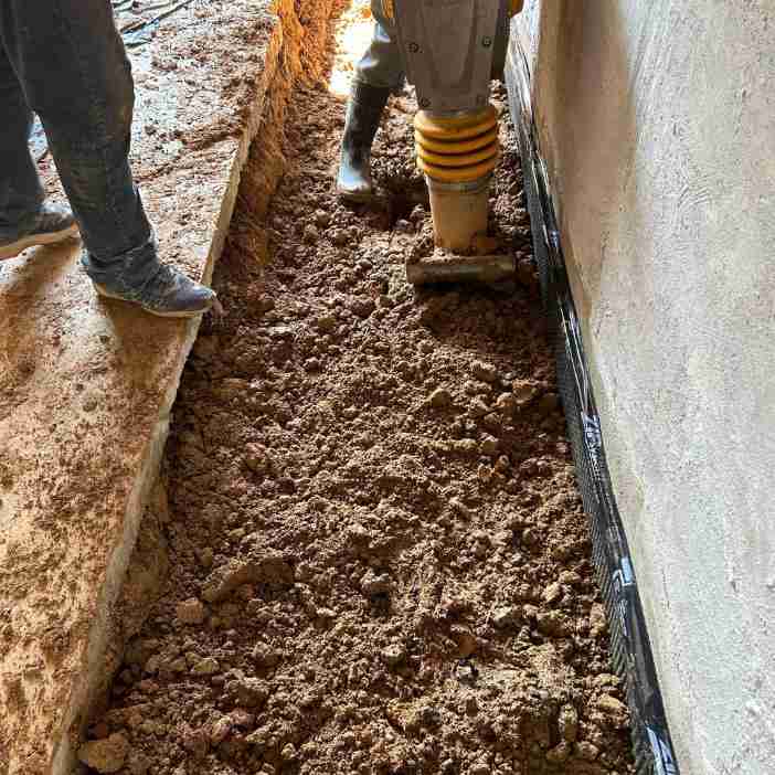 Construction workers compacting dirt with a machine next to a building foundation.