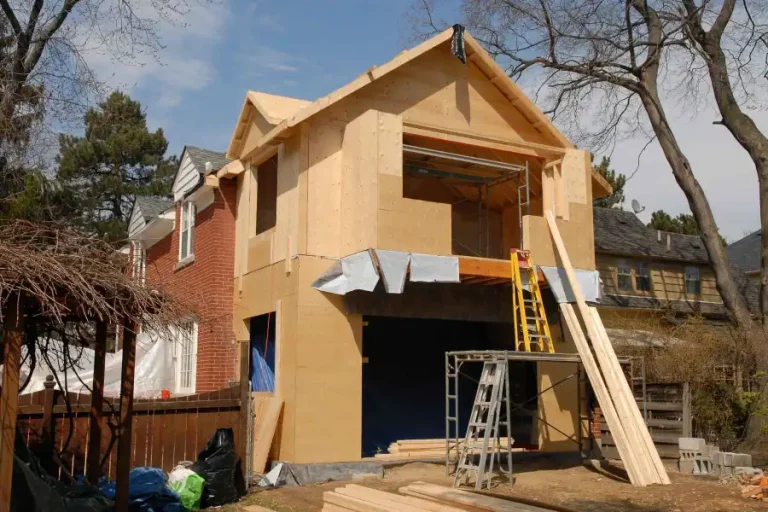 A house under construction with exposed wooden framing and scaffolding, adjacent to an older brick house. A ladder leans against the structure, and building materials are scattered around the site.