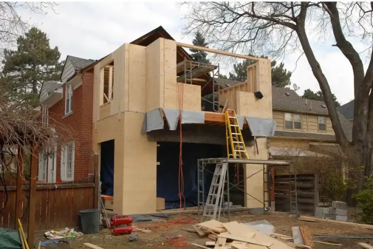 A partially constructed home addition with a visible wooden frame and exposed roof beams. There are ladders, scaffolding, and construction materials scattered around the site. The existing red brick house is seen on the left.