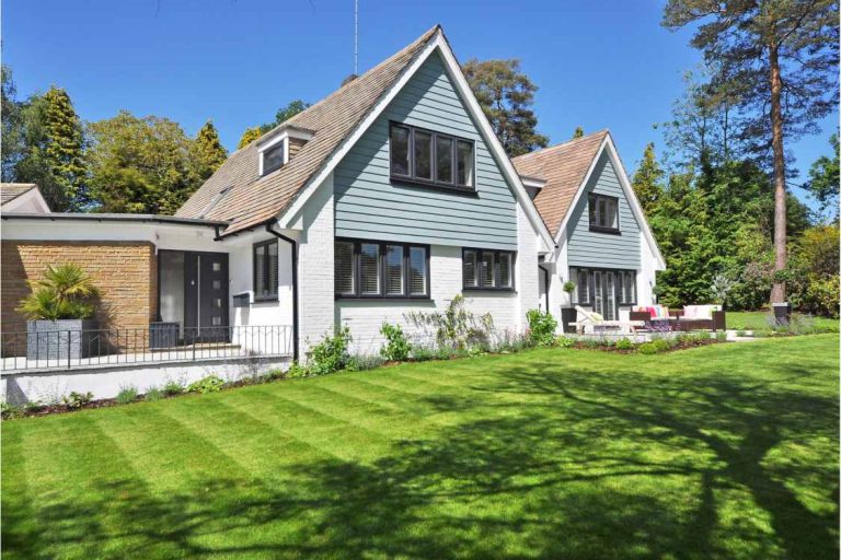 Modern suburban house with sloped roofs, large windows, and a well-maintained front lawn, surrounded by trees.
