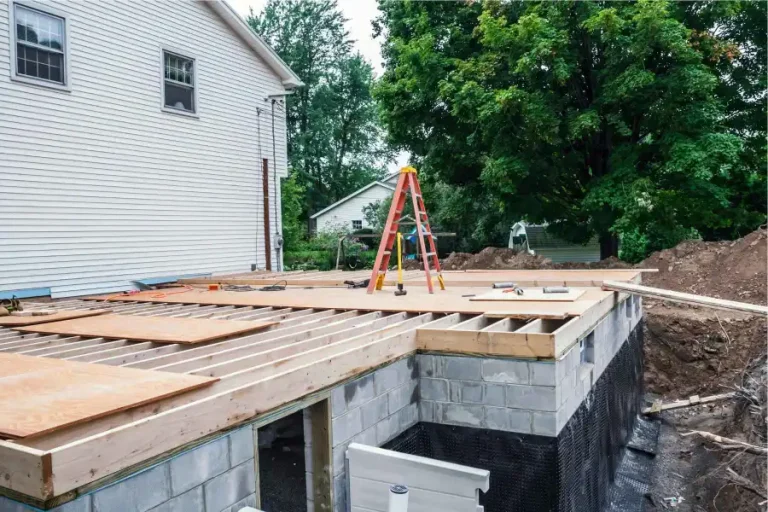 A house under construction with a partially built extension featuring exposed wooden beams and a plywood floor, a ladder and tools on top, surrounded by trees and other homes.