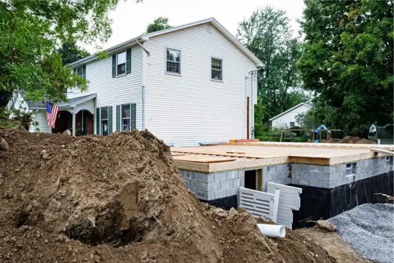 A two-story white house with dark shutters under construction, featuring a new foundation partially covered by wooden boards. Large piles of dirt and construction materials are visible in the yard.