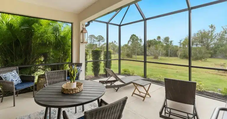 Screened patio with wicker furniture, a round table with a plant centerpiece, and a green outdoor view with trees.