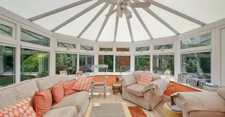A cozy sunroom with a high ceiling, featuring beige sofas adorned with coral and orange cushions, a rocking chair, and large windows overlooking a garden.