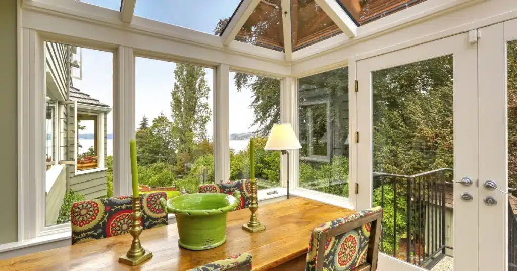 Sunlit dining area with glass walls and ceiling, featuring a wooden table, colorful chairs, decorative green pot, and views of trees and distant water.
