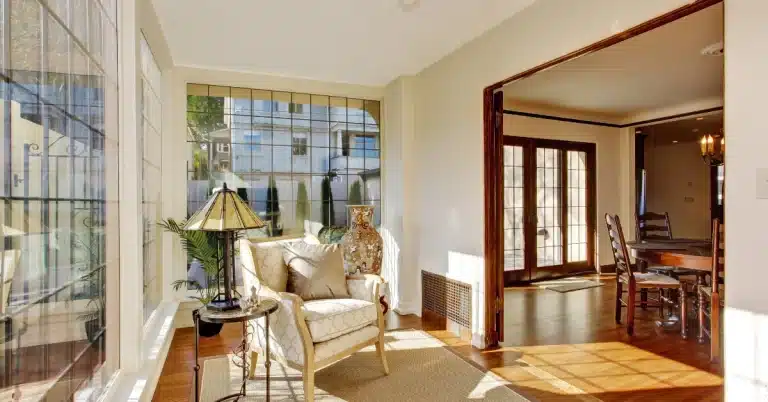 Sunny room with large windows, featuring an armchair, side table, and decorative vase, leading into a dining area with a wooden table and chairs.