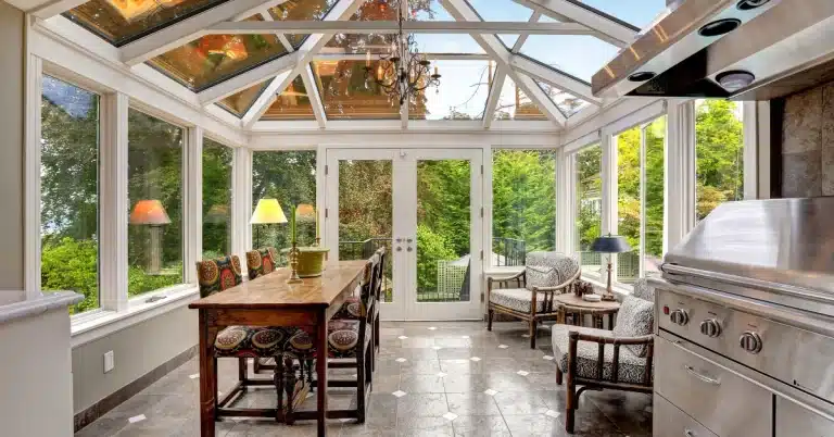 A bright sunroom with a glass ceiling and walls, featuring a wooden dining table with colorful chairs, cushioned armchairs, a stainless steel grill, and lush greenery visible outside.