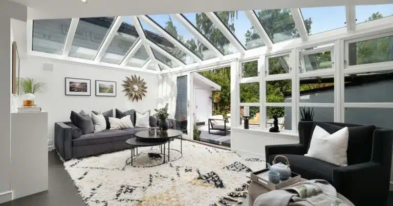 A modern living room with large glass windows and a skylight, featuring a dark gray sofa, a white and black patterned rug, a black armchair, and a coffee table. The room opens to a patio with outdoor seating and lush greenery.