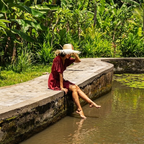 A woman in a red dress and straw hat sits on a curved stone ledge, dipping her feet into a pond surrounded by lush greenery.