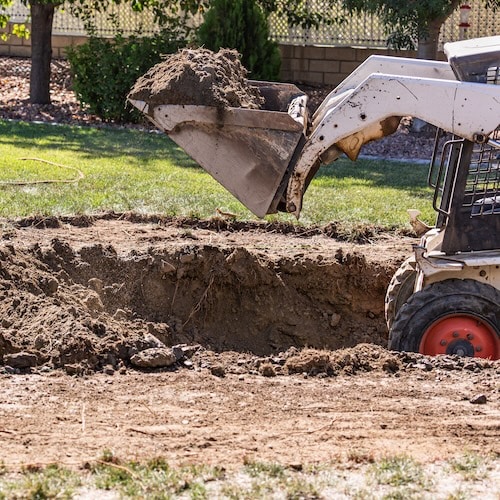 A small excavator scooping up dirt from a large hole in a backyard.