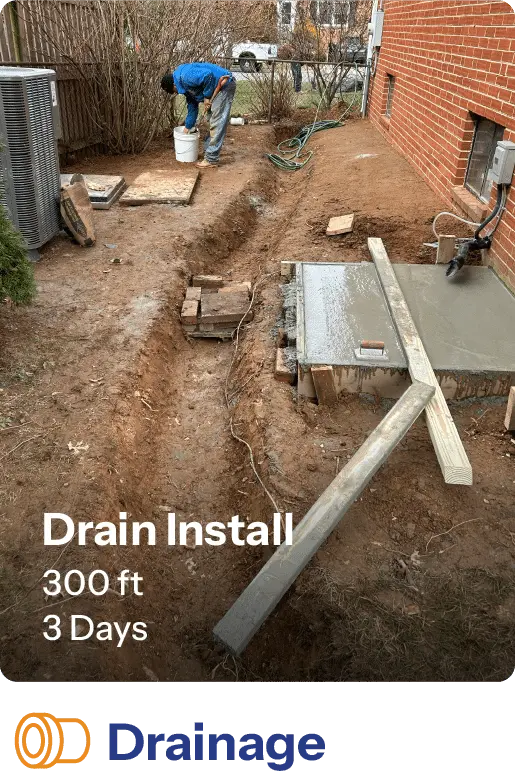 Worker installing a 300-foot drainage system over three days, with a trench visible next to a brick wall and equipment nearby.