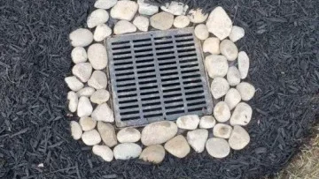 Box drain with metal grate hidden in a mulch bed, bordered by river rocks for drainage and erosion control.
