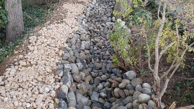 Shallow ditch lined with river rocks to guide rainwater through a backyard and prevent flooding.