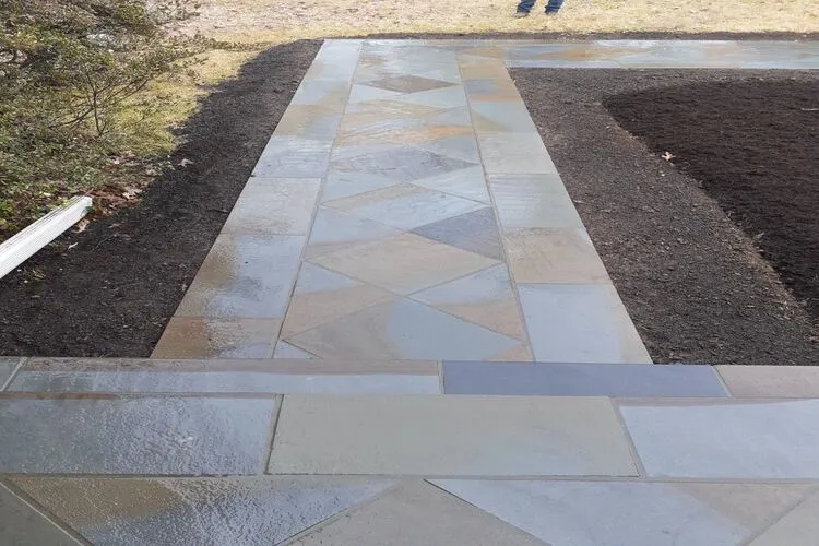 A wet stone pathway with a geometric pattern, surrounded by soil and grass.