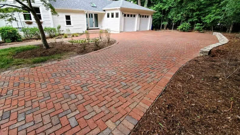 Brick driveway leading to a white house with two garage doors, surrounded by trees.
