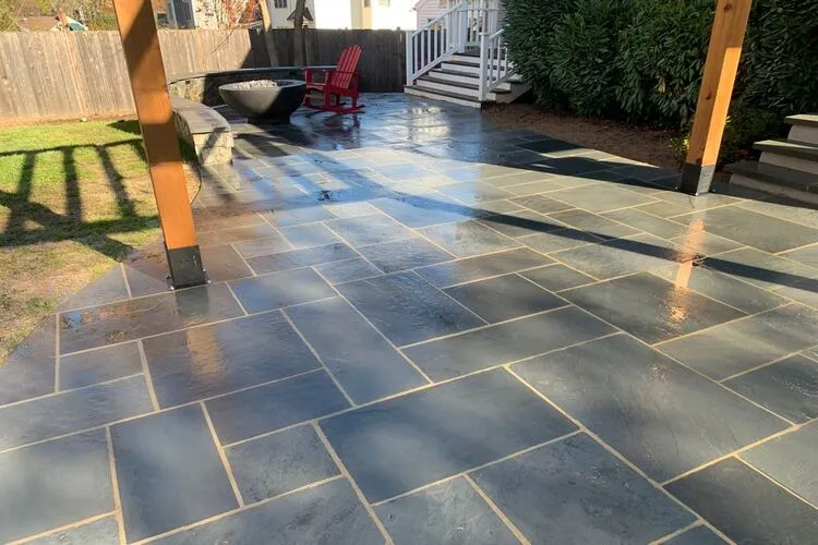 Wet stone patio with red chairs, a fire pit, and wooden pergola in a backyard setting.