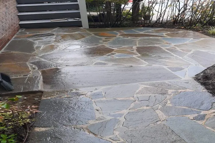 Wet stone patio with irregular shaped tiles near steps and shrubs.