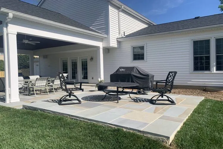 Backyard patio with seating, table, and a covered grill adjacent to a house with white siding.